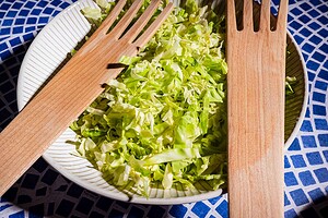 A white bowl filled with cabbage salad, accompanied by two large wooden salad utensils, sits on a blue and white patterned surface.