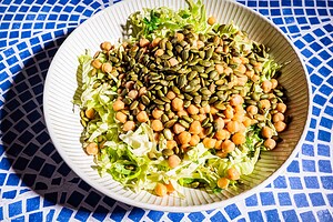 A bowl containing shredded lettuce, chickpeas, pumpkin seeds, and hints of cabbage salad sits on a blue and white mosaic surface.