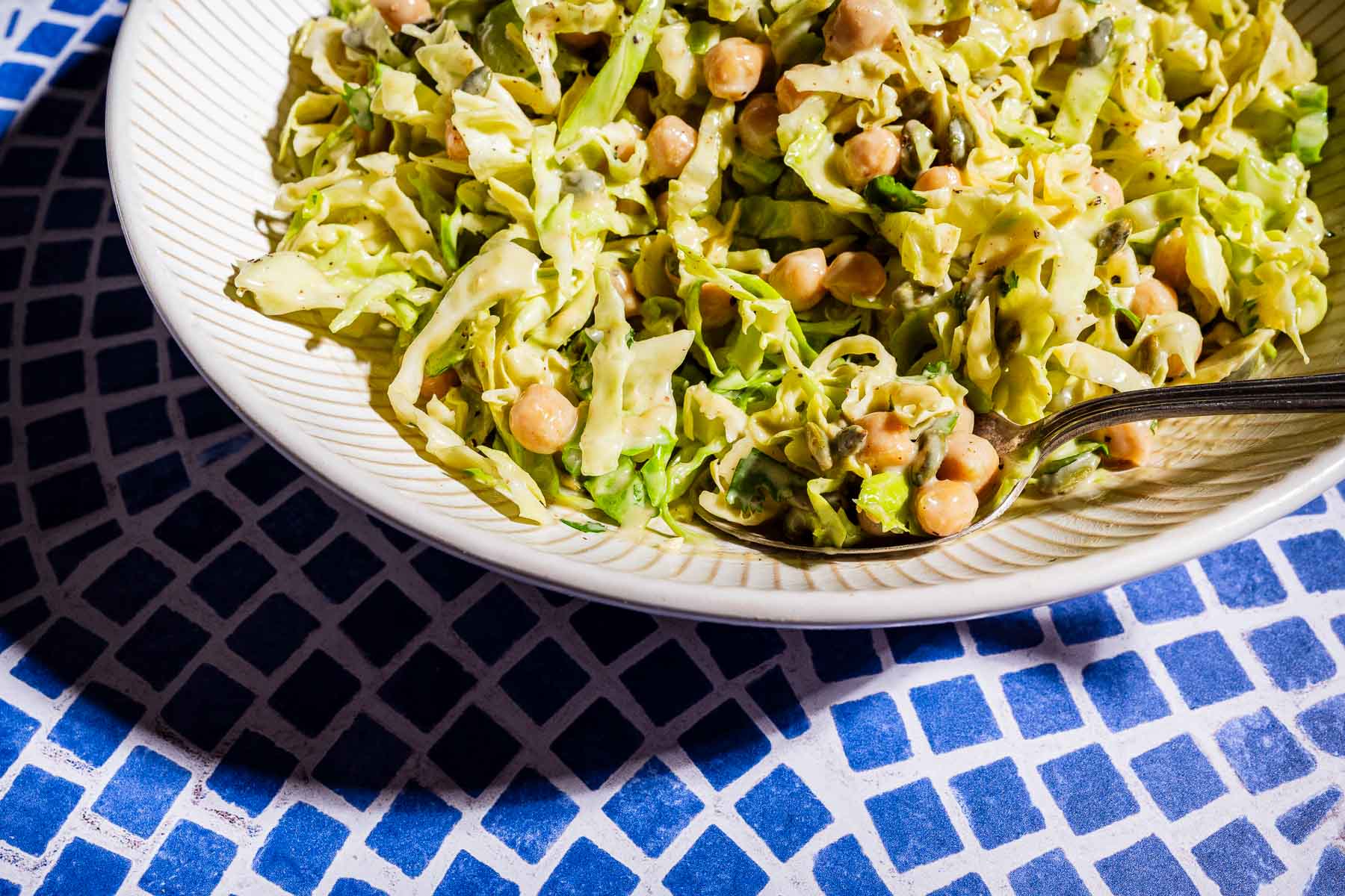 A bowl of fresh cabbage salad with chickpeas and creamy dressing, served with a spoon on a patterned blue and white table.