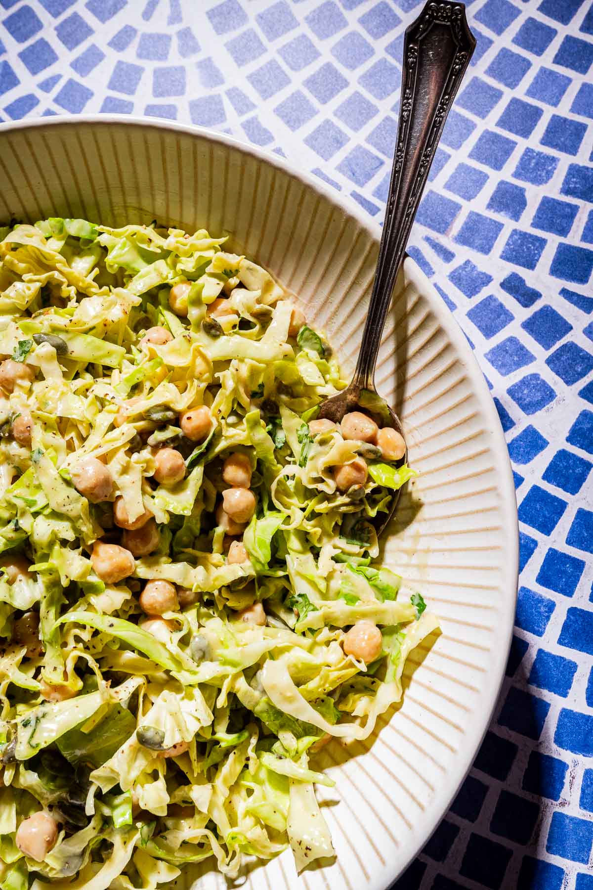 A fresh bowl of cabbage salad with chickpeas and herbs, served with a fork on a blue and white mosaic tile surface.