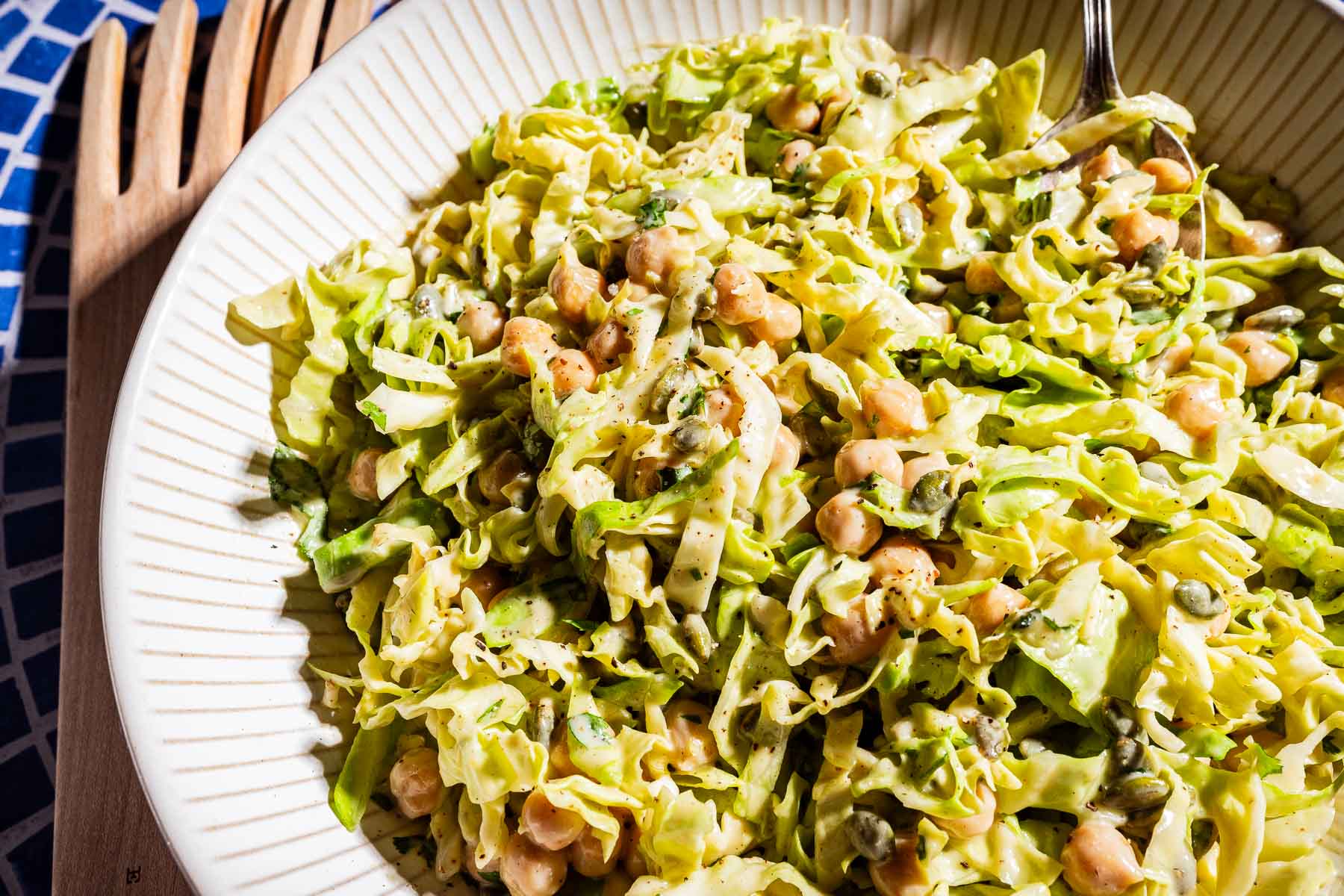 A close-up of a white bowl filled with a fresh cabbage salad featuring shredded lettuce, chickpeas, capers, and a creamy dressing, with a serving fork and spoon beside it.