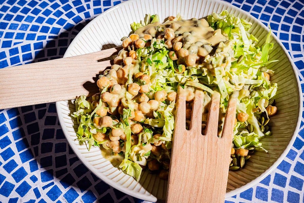 A bowl of chopped lettuce and chickpeas topped with creamy dressing, reminiscent of a fresh cabbage salad, served with wooden salad tongs on a blue patterned surface.