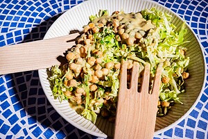 A bowl of chopped lettuce and chickpeas topped with creamy dressing, reminiscent of a fresh cabbage salad, served with wooden salad tongs on a blue patterned surface.