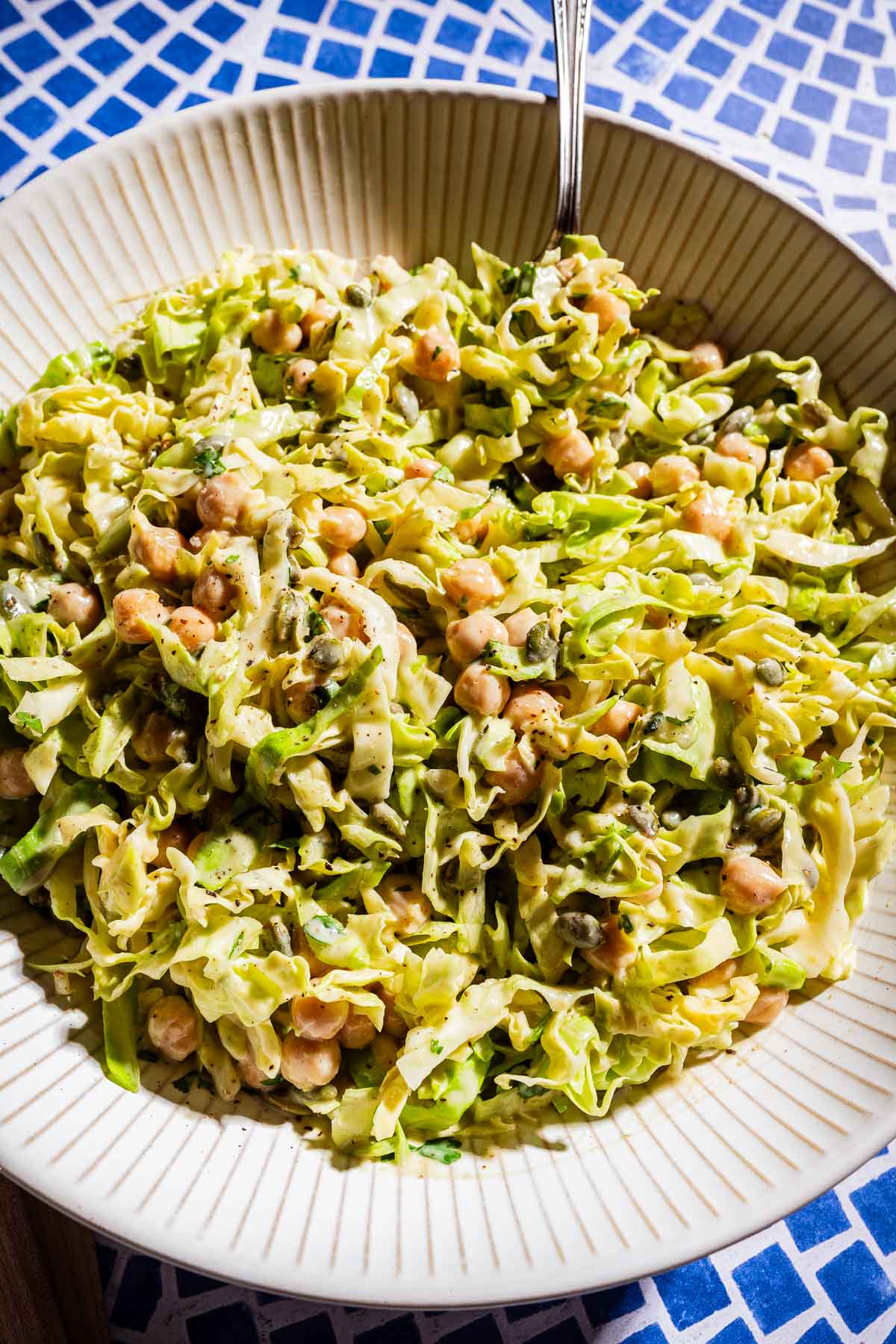 A bowl of vibrant cabbage salad featuring chickpeas, fresh herbs, and a creamy dressing, served with a fork on a blue-and-white tiled surface.