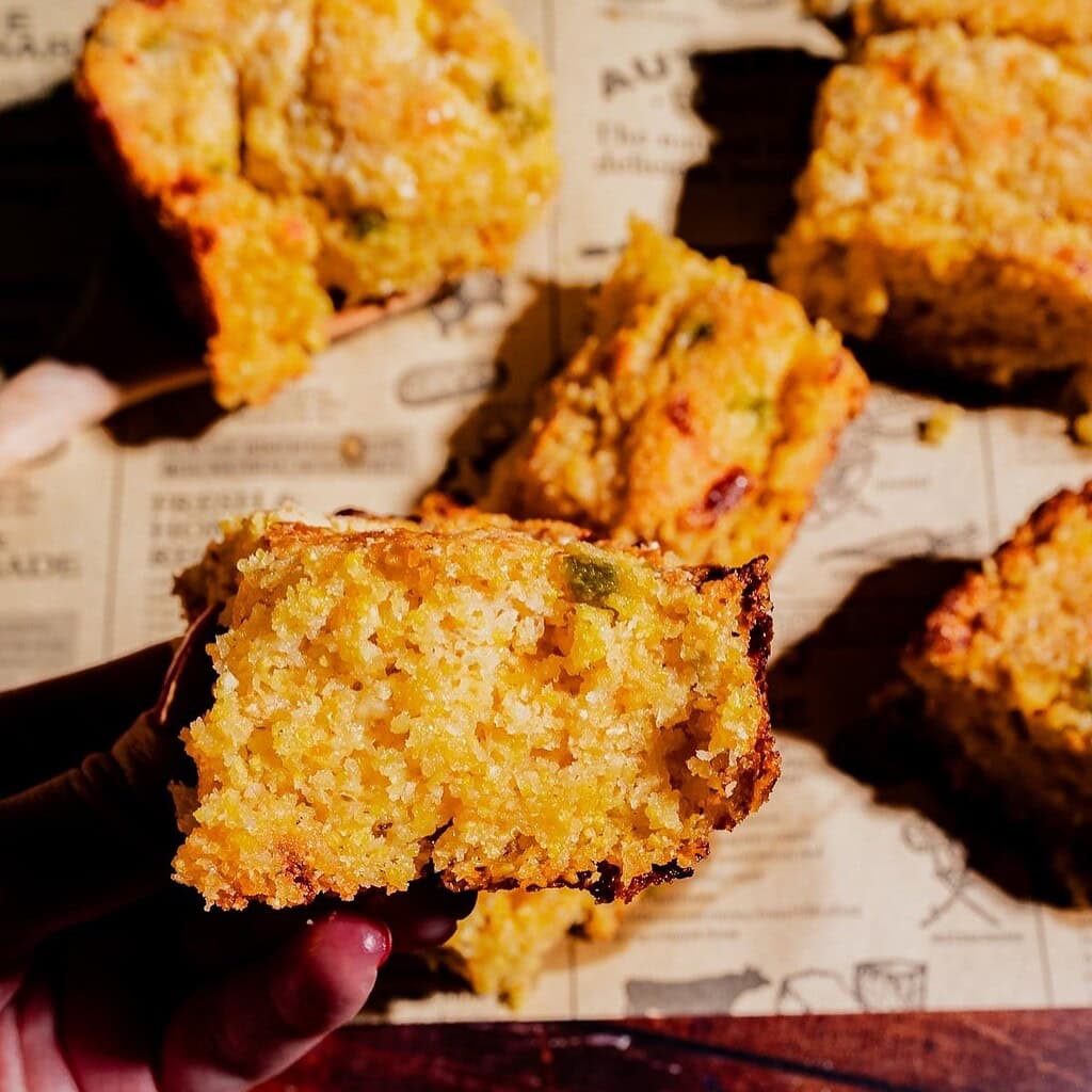 A hand holds a piece of cornbread with visible bits of vegetables; several more pieces are arranged on paper printed with text in the background.