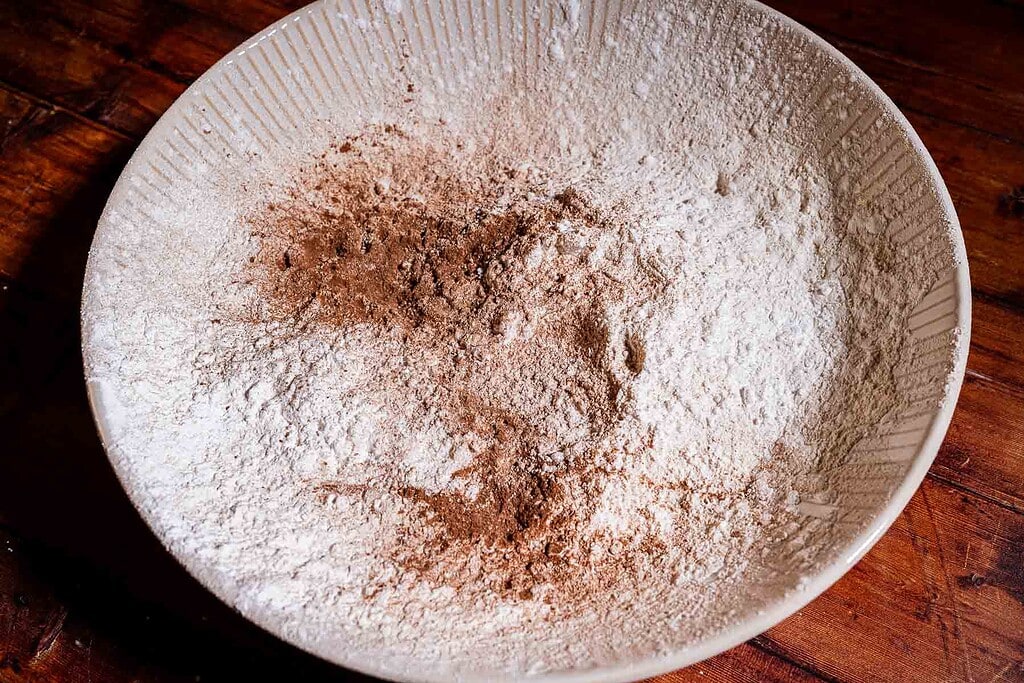 A large ceramic bowl with flour, ground spices, and strawberry buckwheat cake ingredients mixed together sits on a wooden surface.