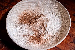 A large ceramic bowl with flour, ground spices, and strawberry buckwheat cake ingredients mixed together sits on a wooden surface.