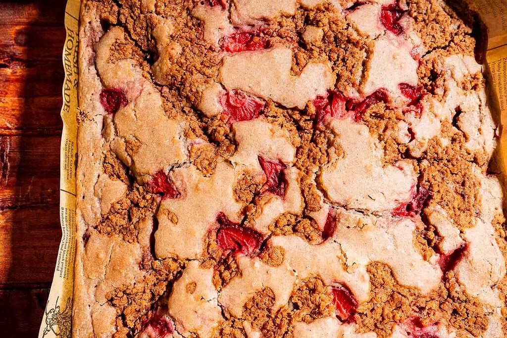 A close-up of a baked strawberry buckwheat cake in a parchment-lined pan, showing a golden brown, crumbly topping with visible strawberry pieces.