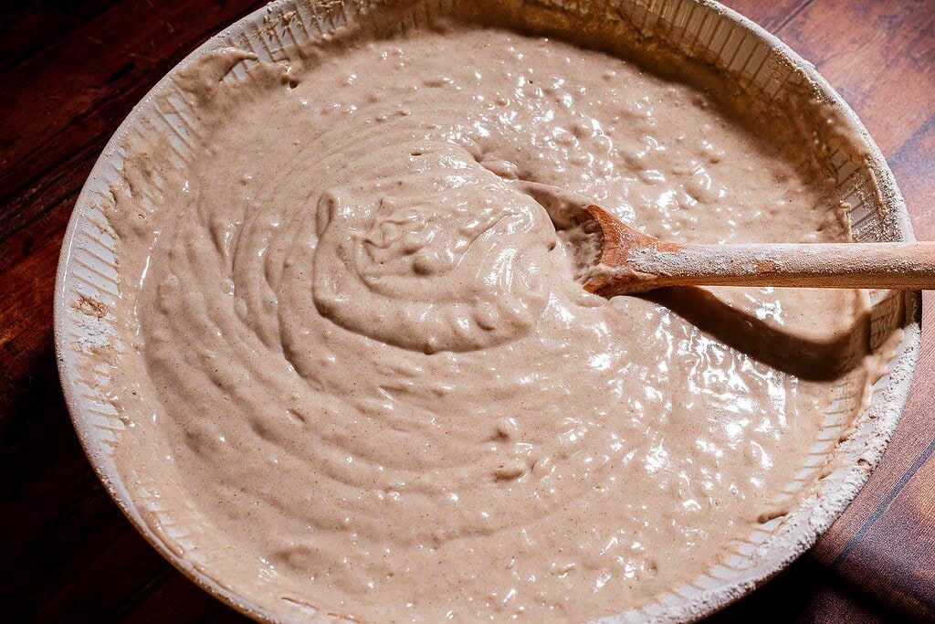 A wooden spoon stirs thick, beige batter for strawberry buckwheat cake in a large ceramic bowl on a wooden surface.