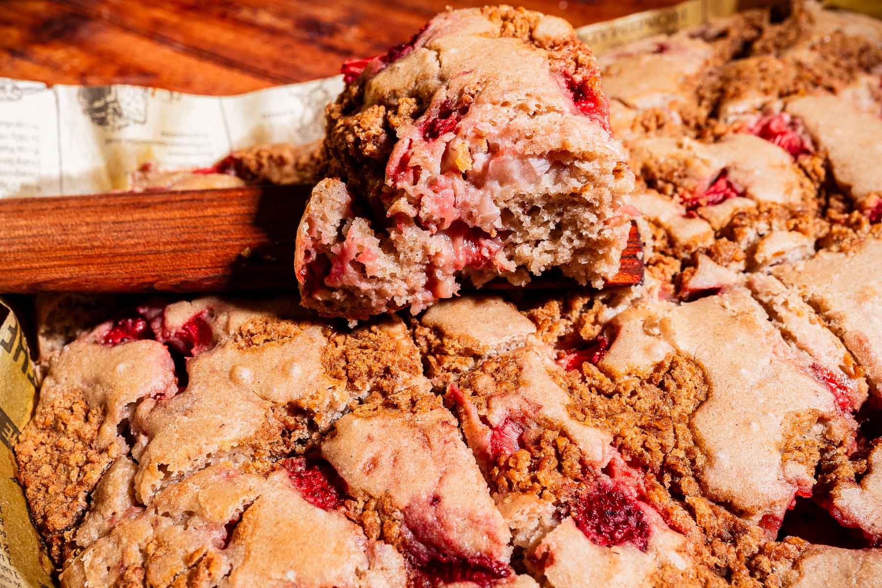 A close-up of a baked strawberry buckwheat cake with a golden-brown crust, cut into squares, with one piece stacked on top of the rest.