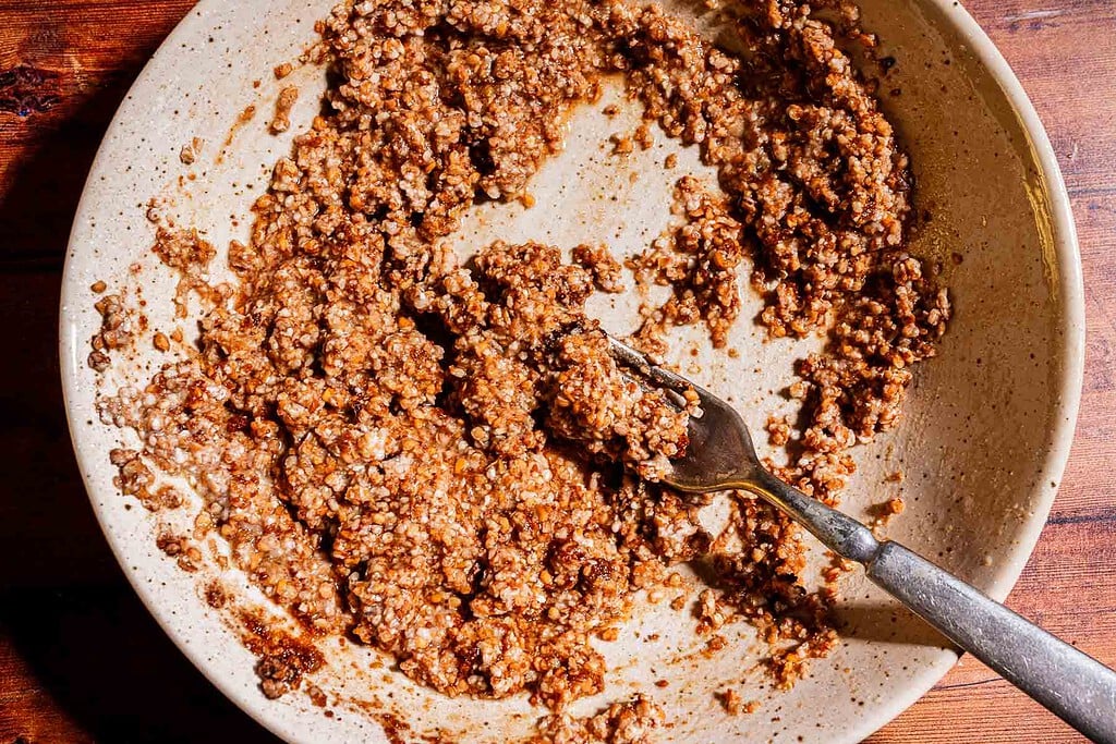 A ceramic bowl contains a mixture of spices and ground meat being mashed with a fork on a wooden surface, next to a freshly baked strawberry buckwheat cake.