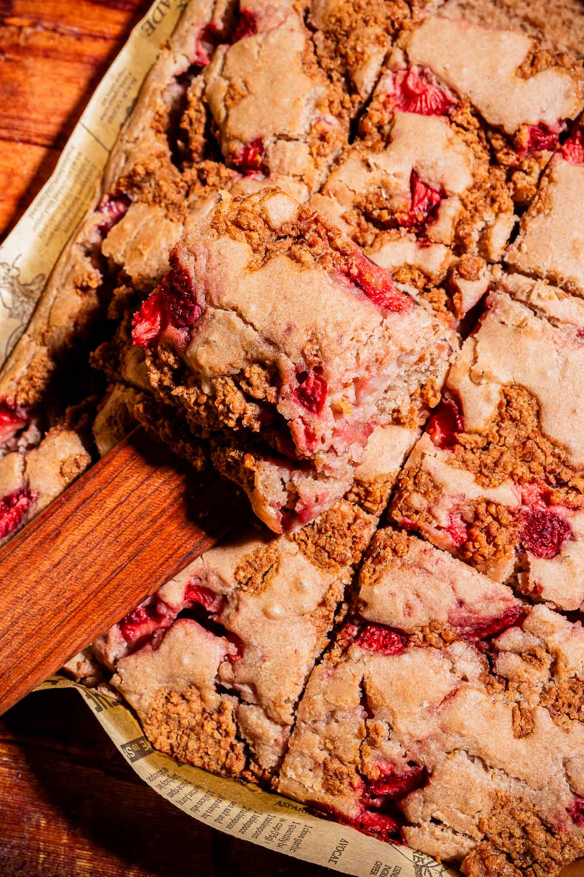 A close-up of strawberry crumb bars cut into squares, with one bar being lifted on a wooden spatula, reminiscent of a classic strawberry buckwheat cake.