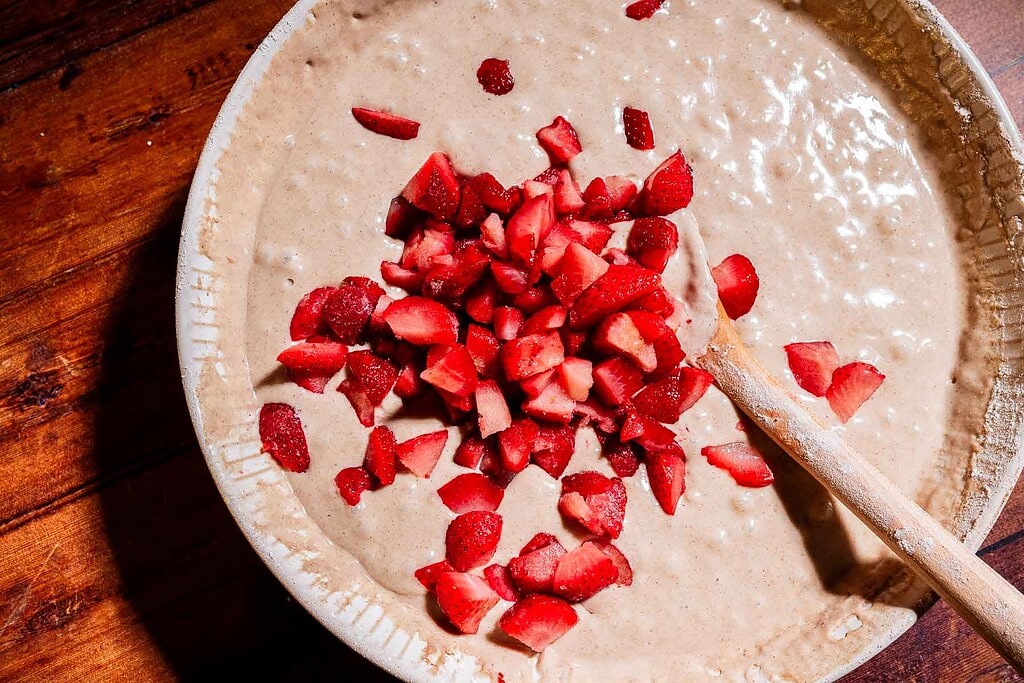 A mixing bowl of batter for strawberry buckwheat cake with chopped strawberries and a wooden spoon on a wooden surface.