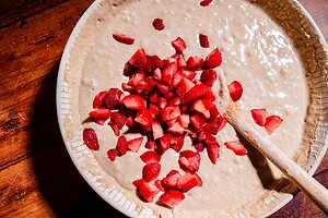 A mixing bowl of batter for strawberry buckwheat cake with chopped strawberries and a wooden spoon on a wooden surface.