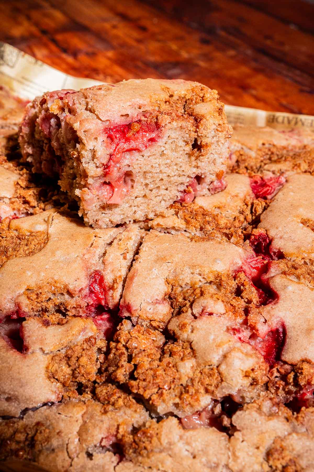 A close-up of sliced strawberry buckwheat cake with a crumb topping, showing a piece resting on top of the rest of the cake on a wooden surface.
