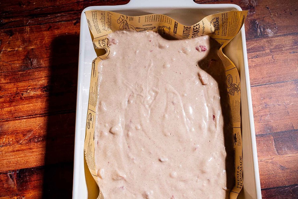 A baking dish lined with printed parchment paper filled with uncooked strawberry buckwheat cake batter sits on a wooden surface.