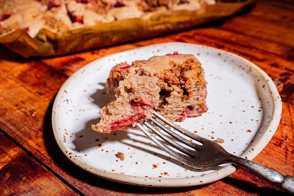 A slice of strawberry buckwheat cake with a bite taken out sits on a speckled white plate with a fork, on a wooden table. More cake is visible in the background.