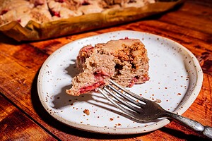 A slice of strawberry buckwheat cake with a bite taken out sits on a speckled white plate with a fork, on a wooden table. More cake is visible in the background.