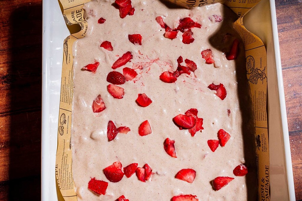 A baking dish lined with parchment paper contains strawberry buckwheat cake batter, generously topped with pieces of fresh strawberries, ready to be baked.