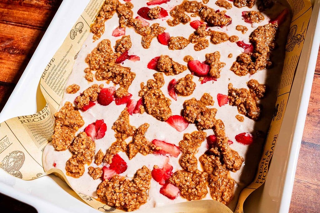 A baking dish lined with parchment paper holds an unbaked strawberry buckwheat cake mixture topped with chopped strawberries and brown oat crumble.