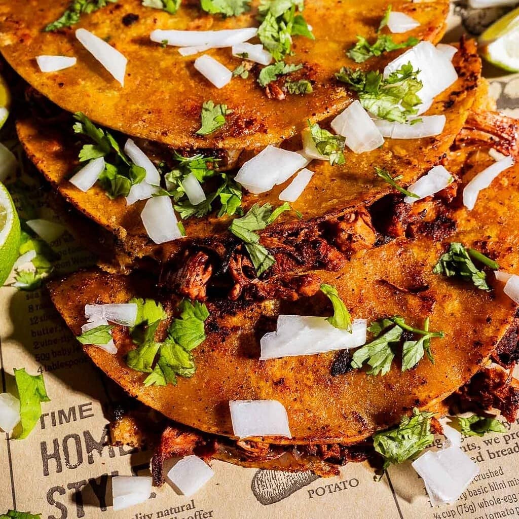 Three jackfruit birria tacos filled with shredded goodness, topped with chopped onions and cilantro, served on newspaper with lime wedges on the side.