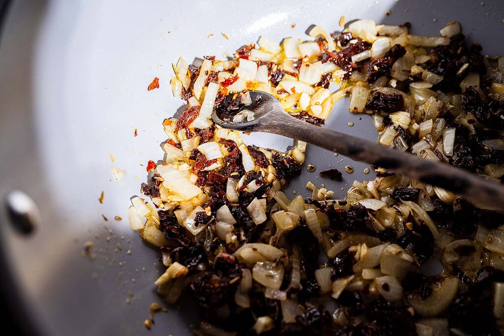 Chopped onions and sun-dried tomatoes sautéing in a pan with a wooden spoon, creating the irresistible base for marry me lasagna soup.