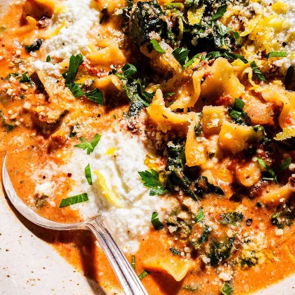 Close-up of creamy marry me lasagna soup with pasta, spinach, tomato-based broth, ricotta cheese, grated parmesan, fresh parsley, and a spoon on the side.