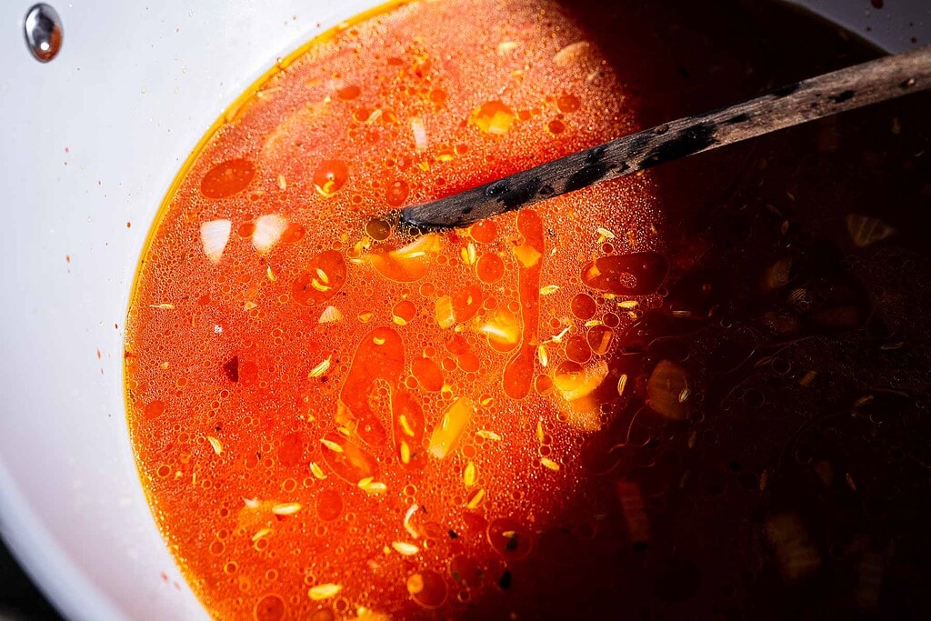 A close-up of red marry me lasagna soup with oil, spices, and chopped onions in a pot, with a wooden spoon resting on the surface.