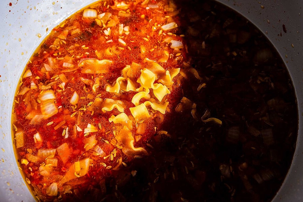 A close-up of marry me lasagna soup with curly noodles, chopped onions, and a rich red broth in a white bowl, partially covered in shadow.