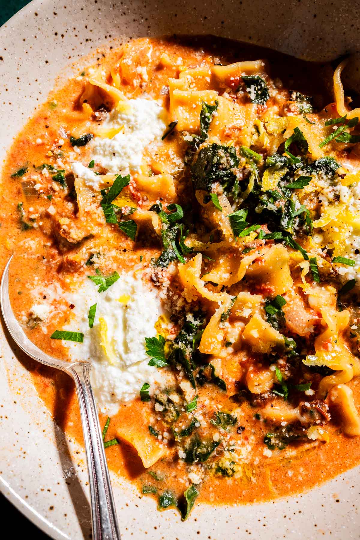A bowl of creamy marry me lasagna soup with pasta, spinach, tomato sauce, ricotta cheese, grated cheese, and fresh herbs, served with a spoon.