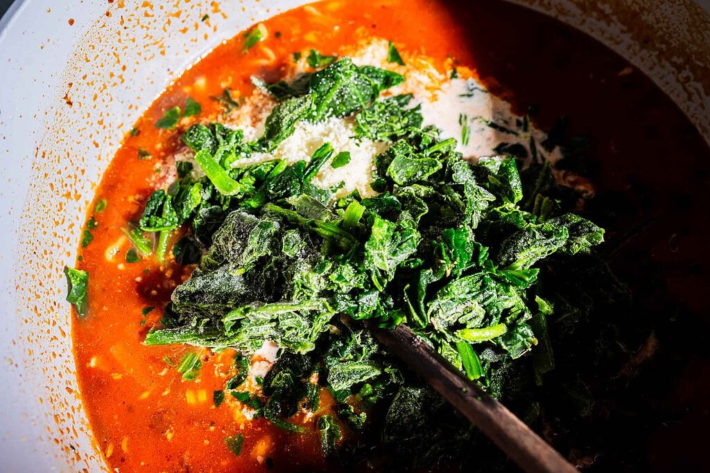 A close-up of frozen spinach being added to a pot of tomato-based marry me lasagna soup with visible seasonings and a wooden spoon.