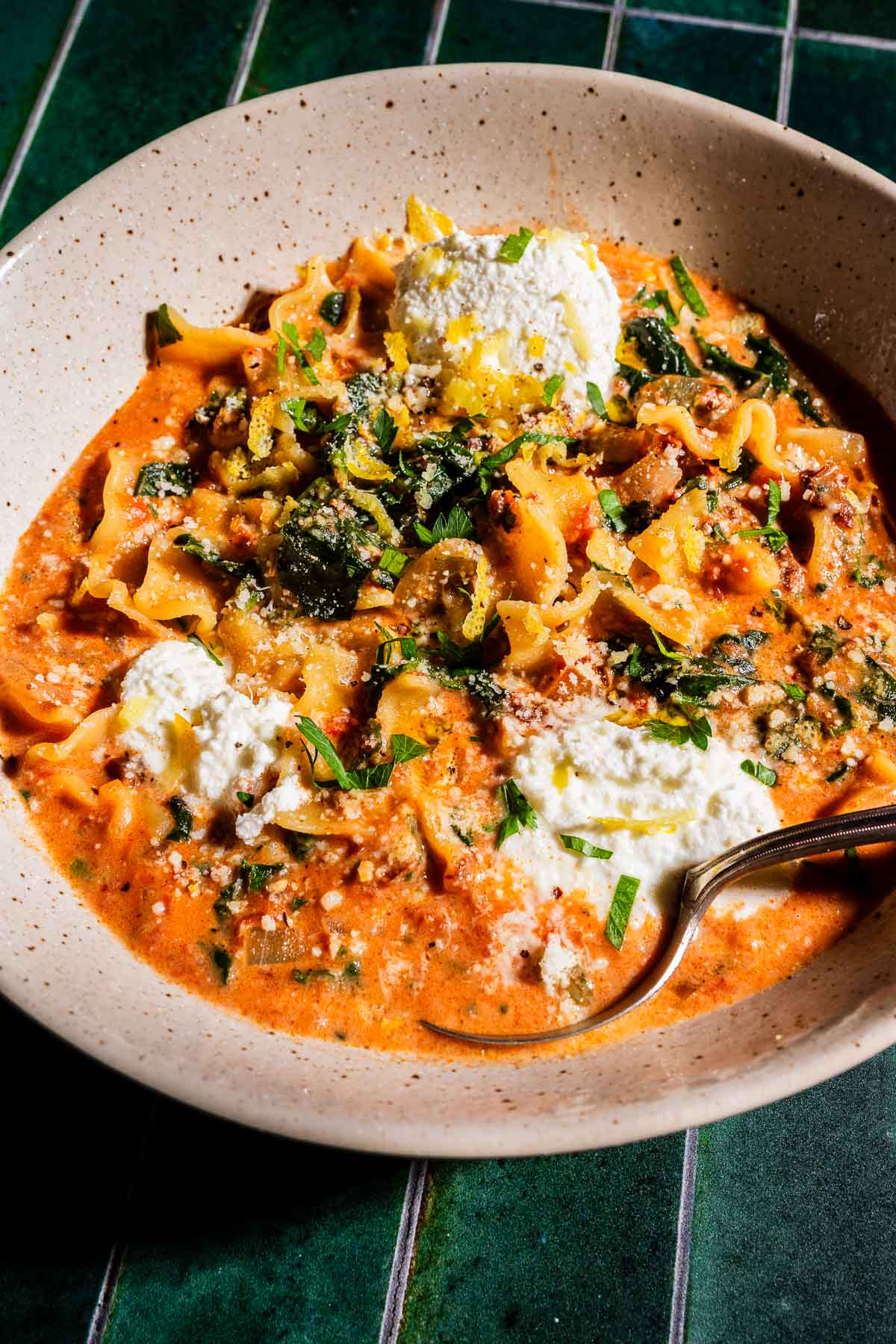 A bowl of marry me lasagna soup topped with dollops of ricotta cheese, grated parmesan, and chopped herbs, with a spoon resting inside.