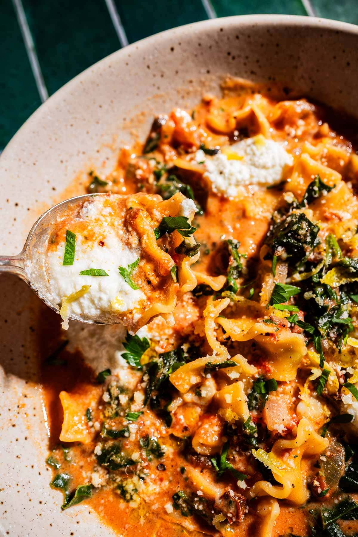A close-up of a bowl of marry me lasagna soup with noodles, tomato sauce, cheese, and herbs, as a spoon lifts a hearty portion from the bowl.