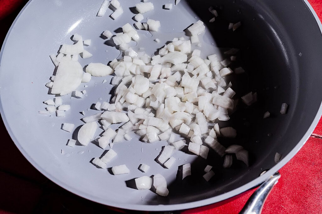 Chopped white onions in a gray frying pan, placed on a red surface, ready to be added to a flavorful purple sweet potato curry.