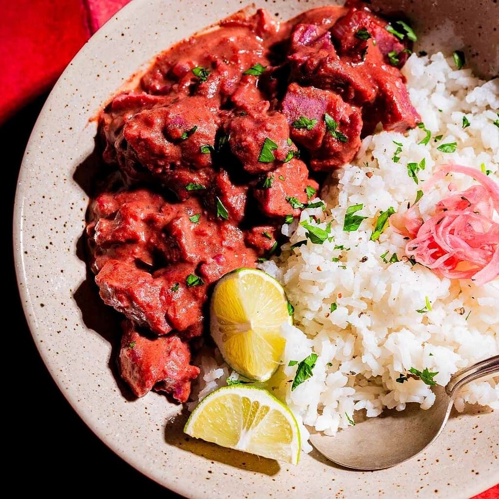 A plate with white rice, beef stew in red sauce, pickled onions, lime wedges, and a spoon, garnished with chopped herbs—served alongside vibrant purple sweet potato curry for a colorful and flavorful meal.