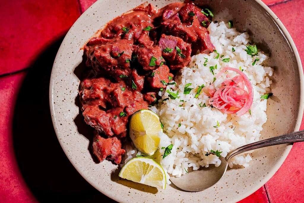 A bowl of white rice topped with pickled onions and chopped herbs, served with a vibrant purple sweet potato curry and lemon wedges, with a spoon on the side.