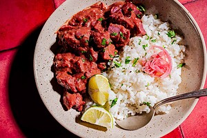 A bowl of white rice topped with pickled onions and chopped herbs, served with a vibrant purple sweet potato curry and lemon wedges, with a spoon on the side.