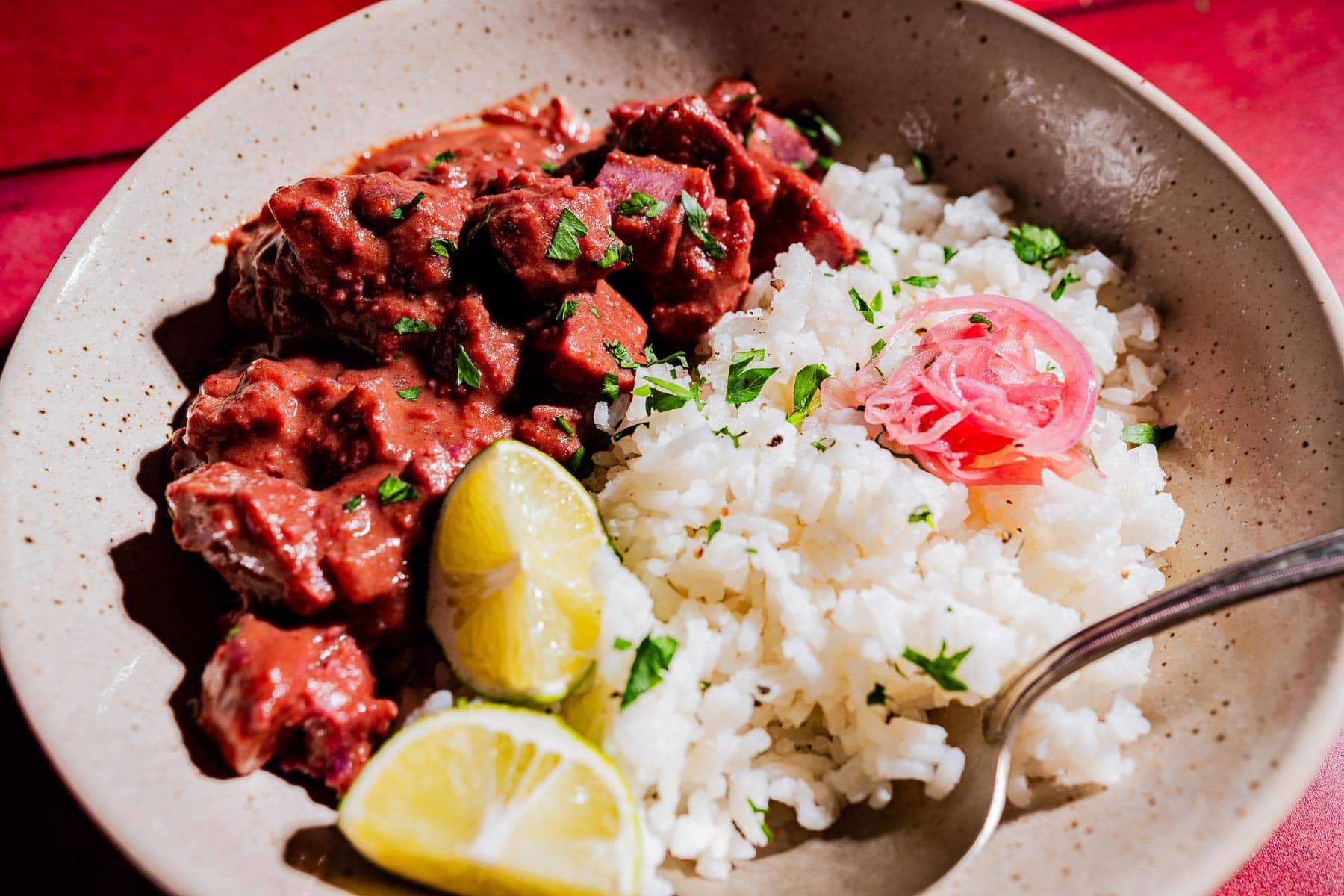 A plate of white rice garnished with herbs and pickled onions, served with beef stew in red sauce, purple sweet potato curry, and lemon wedges, with a spoon on the side.