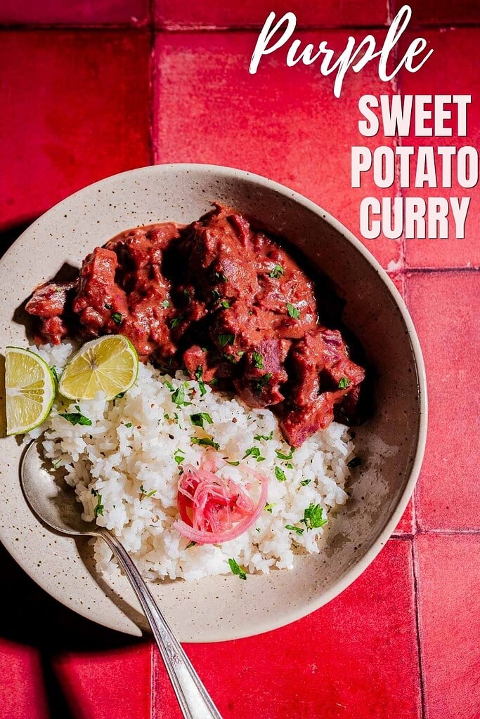 A bowl of white rice and vibrant purple sweet potato curry is garnished with lime wedges, pickled onions, and fresh herbs, set on a bold red tiled surface.