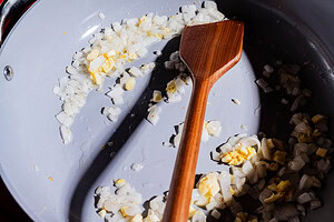 Chopped onions are being sautéed in a light-colored pan with a wooden spatula, creating the aromatic base for a flavorful purple sweet potato curry.