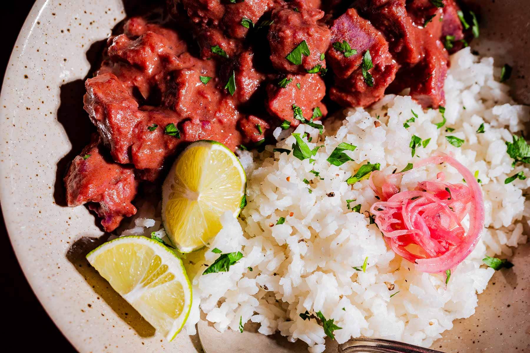 A plate with white rice garnished with cilantro and pickled onions, served alongside a beef stew in red sauce, purple sweet potato curry, and lime wedges.