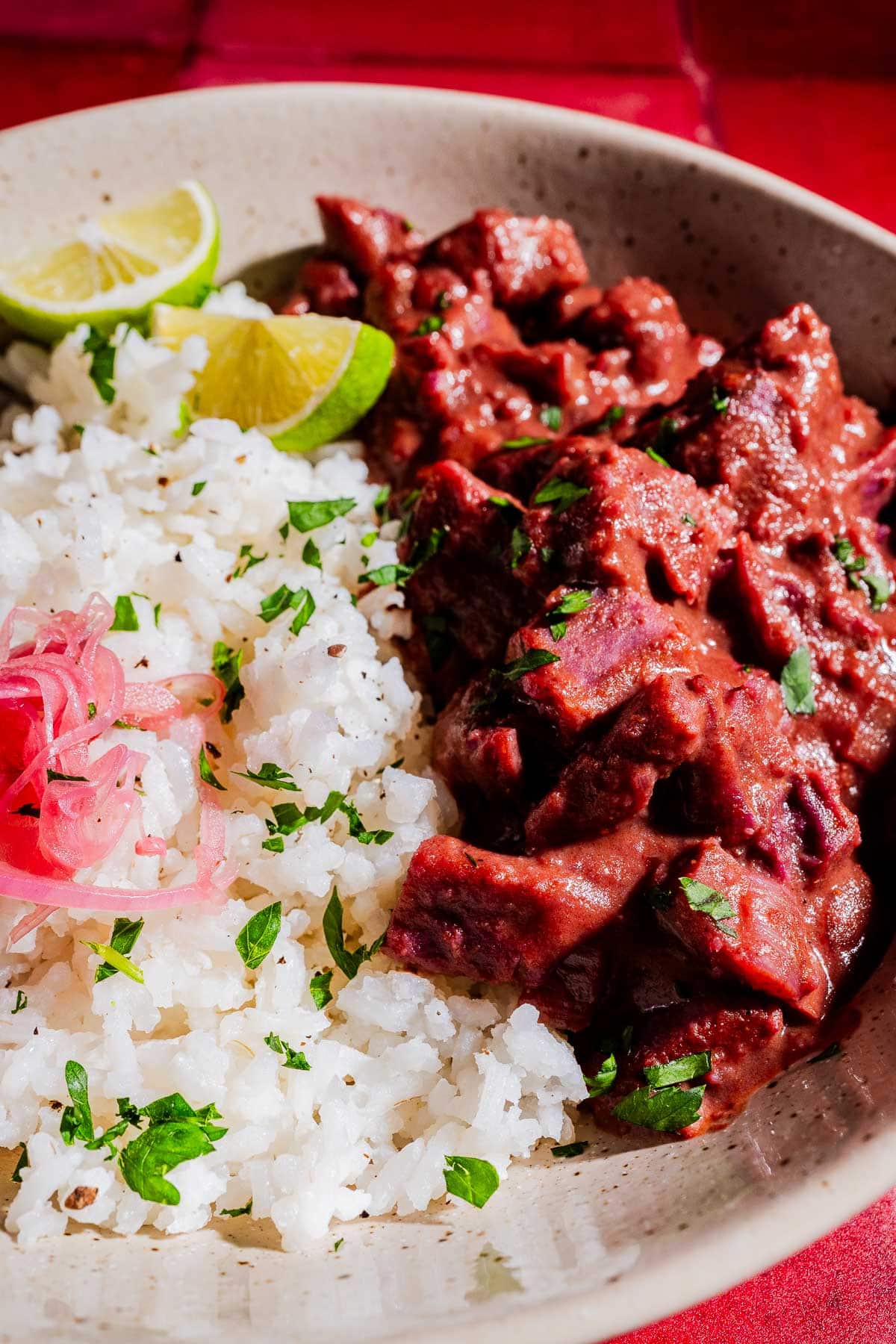 A bowl of white rice garnished with chopped herbs and pickled onions, served with a portion of beef stew in dark red sauce and lime wedges, alongside vibrant purple sweet potato curry.