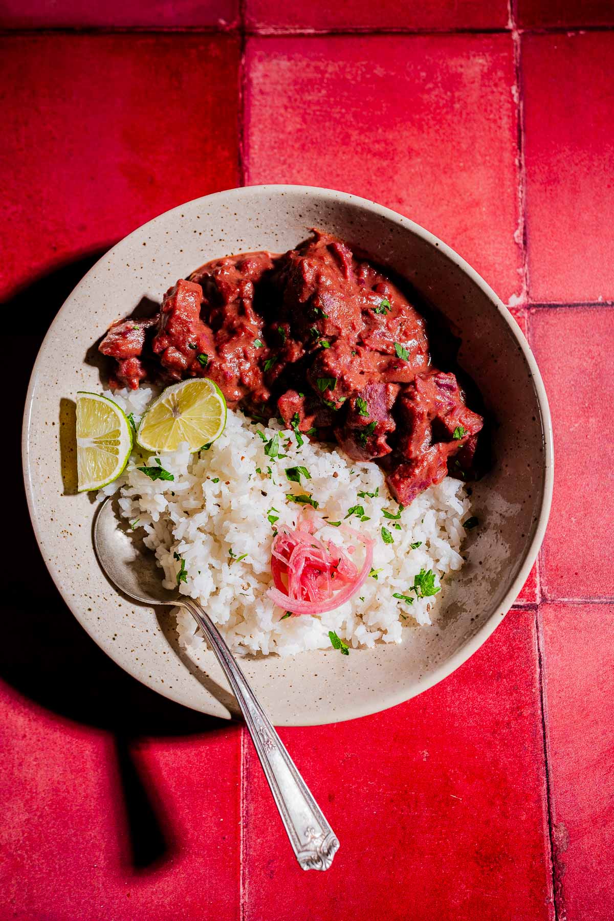 A bowl of white rice with stewed meat in red sauce, purple sweet potato curry, garnished with cilantro, pickled onions, and lime wedges, on a red tiled surface. A spoon rests in the bowl.