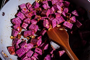 Cubed purple sweet potatoes with spices and onions are being sautéed in a pan with a wooden spoon, starting the base for a flavorful purple sweet potato curry.