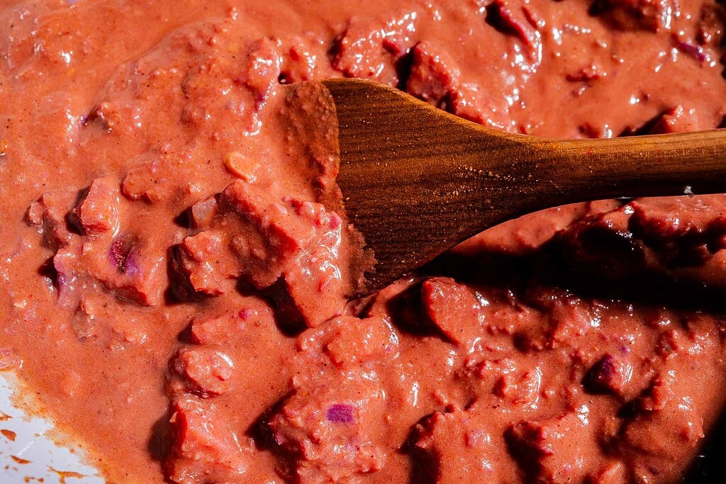 Close-up of a wooden spoon stirring a thick, chunky reddish-brown stew or sauce with visible pieces of meat and beans, reminiscent of a hearty purple sweet potato curry.