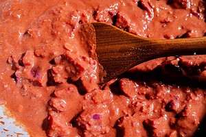 Close-up of a wooden spoon stirring a thick, chunky reddish-brown stew or sauce with visible pieces of meat and beans, reminiscent of a hearty purple sweet potato curry.