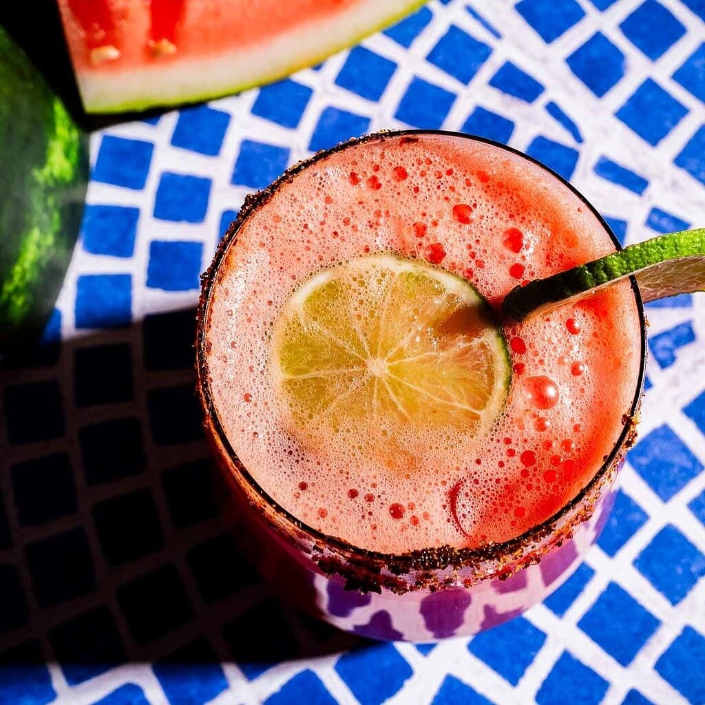 A glass of pink watermelon agua fresca with a lime slice and chili powder on the rim, placed on a blue and white patterned surface.