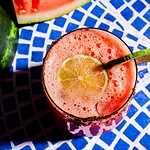 A glass of pink watermelon agua fresca with a lime slice and chili powder on the rim, placed on a blue and white patterned surface.