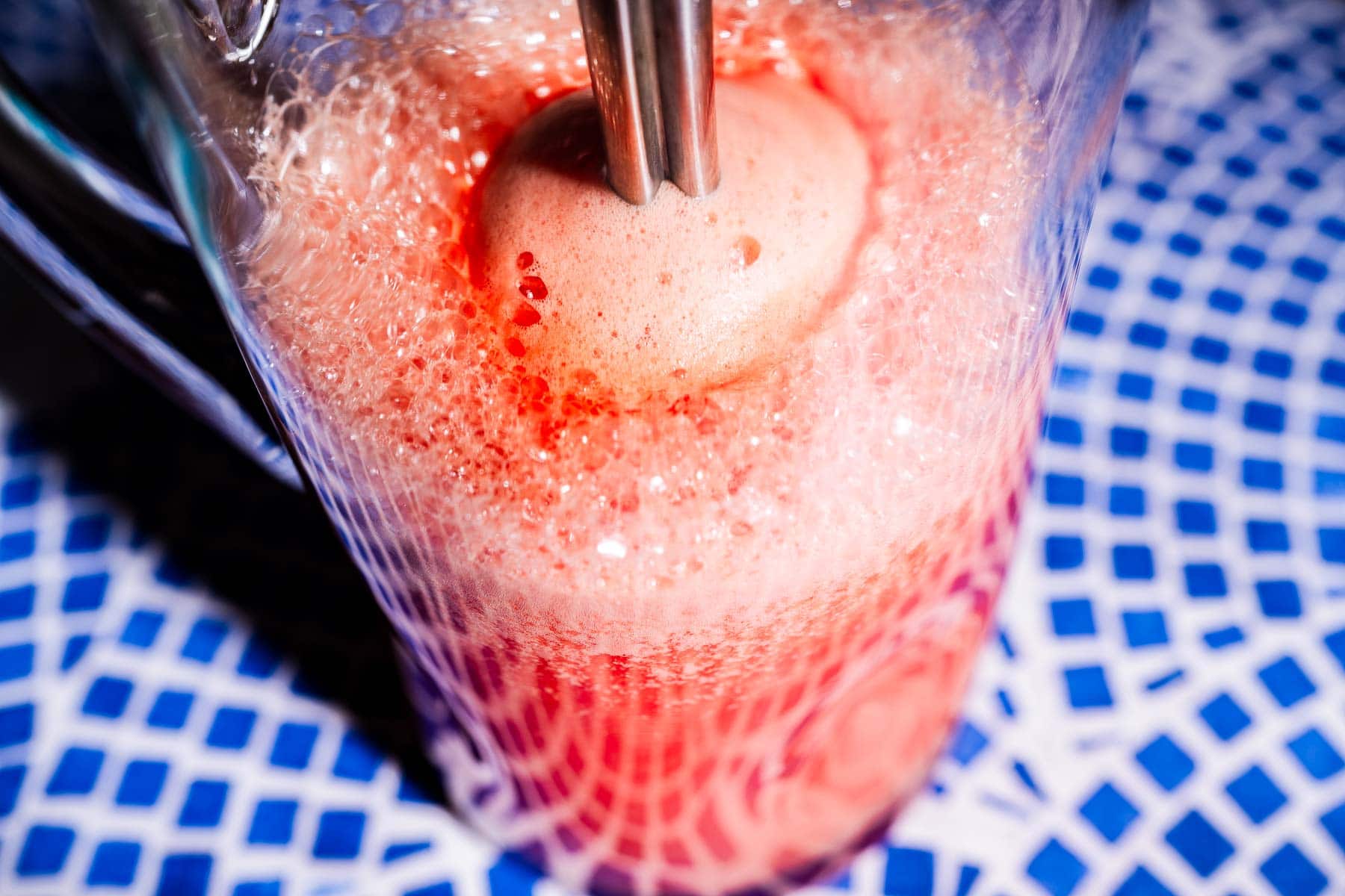 A close-up of a handheld blender mixing frothy watermelon agua fresca in a glass pitcher on a blue patterned surface.