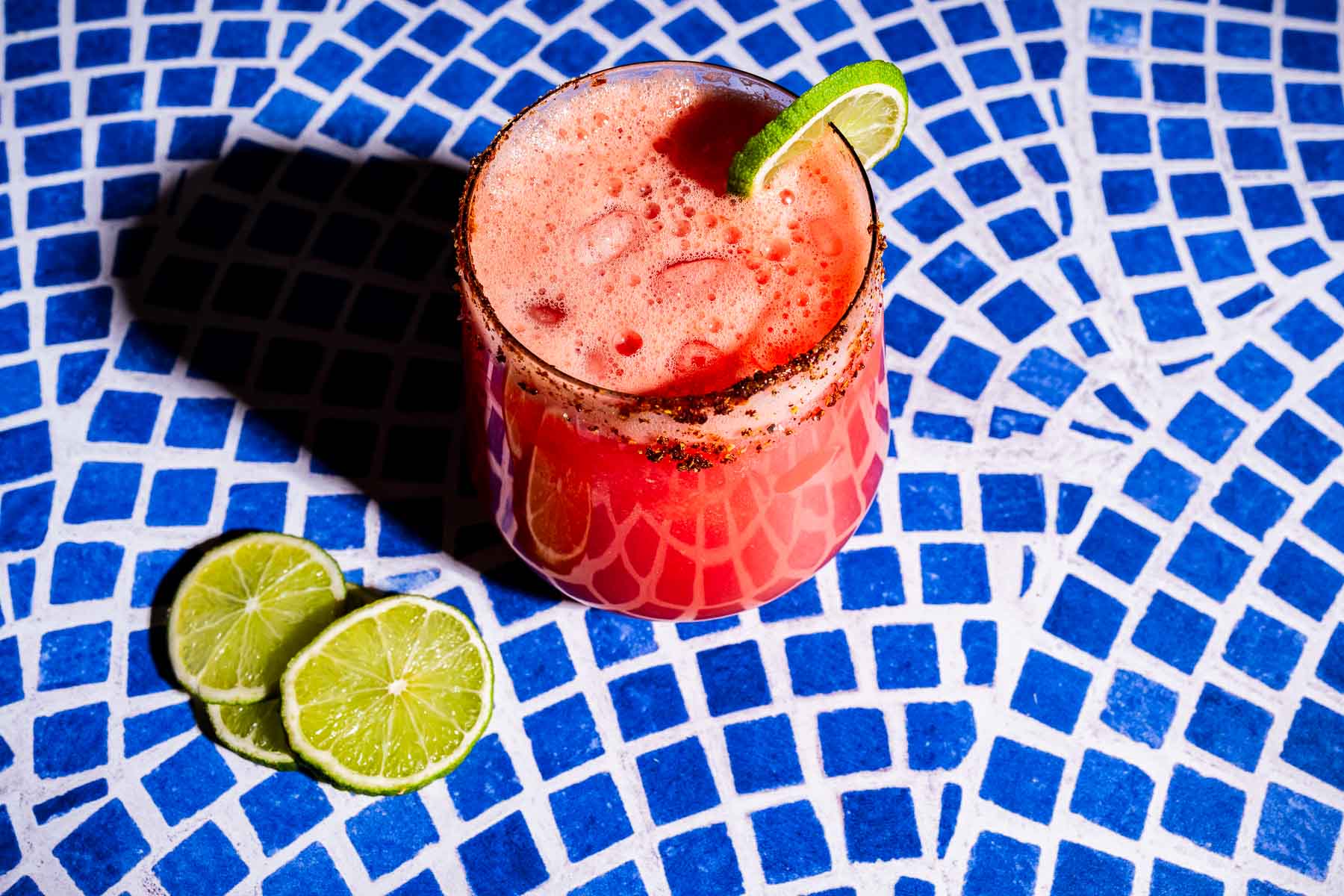 A glass of red watermelon agua fresca cocktail with a chili-rimmed edge and a lime wedge garnish sits on a blue mosaic surface, with two lime slices placed beside the glass.